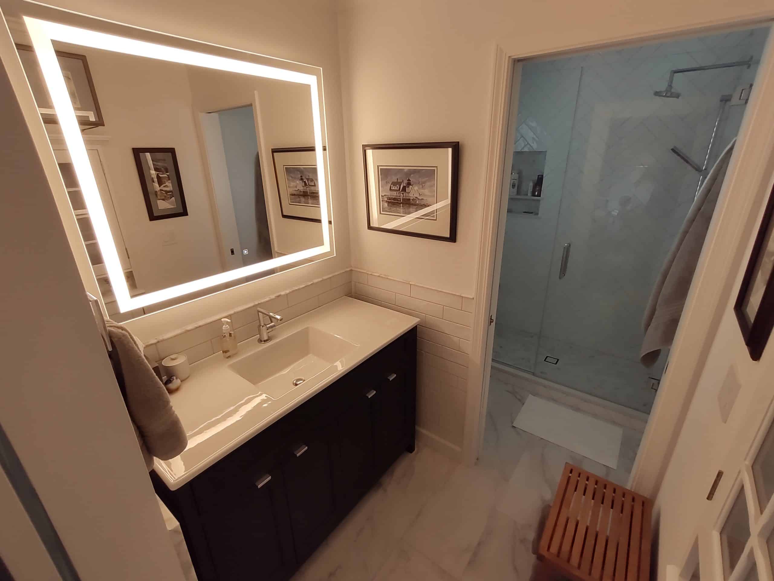 Wide-angle image of a remodeled bathroom with herringbone tile shower. On the left is a vanity with an LED mirror and tiled wall surround.